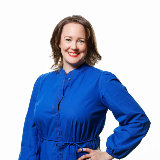Leende kvinna i blått står med vänster hand på höger höft.